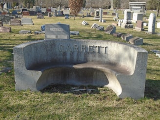 A cemetery scene with numerous headstones of varying shapes and sizes scattered across a grassy area. In the foreground, a curved stone bench is prominently displayed with the name 'Garrett' engraved on it. In the background, more headstones can be seen, along with trees and a clear blue sky.