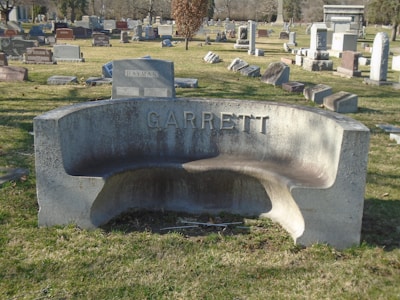 A cemetery scene with numerous headstones of varying shapes and sizes scattered across a grassy area. In the foreground, a curved stone bench is prominently displayed with the name 'Garrett' engraved on it. In the background, more headstones can be seen, along with trees and a clear blue sky.