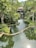A serene environment featuring a winding water body surrounded by lush green vegetation and trees. There is a building at the far end partially hidden by the foliage, suggesting a peaceful retreat in a natural setting.