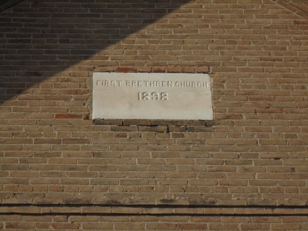 A historic brick wall features an engraved stone plaque. The plaque is inscribed with 'First Brethren Church 1898,' indicating the building's association and the year of its establishment. The bricks are uniformly arranged and vary slightly in color, giving a vintage appearance.