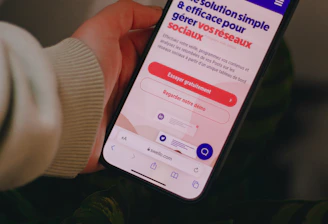 A person holds a smartphone that displays a website in French offering a simple and effective solution for managing social networks. The smartphone screen shows vibrant colors including purple and red text, and the background features green leaves.