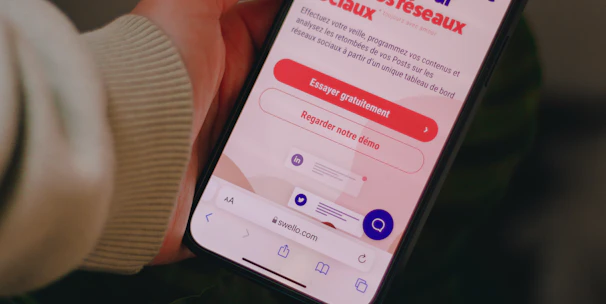 A person holds a smartphone that displays a website in French offering a simple and effective solution for managing social networks. The smartphone screen shows vibrant colors including purple and red text, and the background features green leaves.