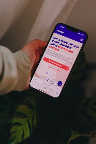 A person holds a smartphone that displays a website in French offering a simple and effective solution for managing social networks. The smartphone screen shows vibrant colors including purple and red text, and the background features green leaves.