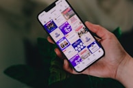 A hand holding a smartphone displaying an Instagram profile grid. The screen features various posts with vibrant colors and text, focusing on motivational content and social media strategies. The background includes leafy plants, adding a natural touch.