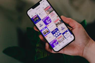 A hand holding a smartphone displaying an Instagram profile grid. The screen features various posts with vibrant colors and text, focusing on motivational content and social media strategies. The background includes leafy plants, adding a natural touch.