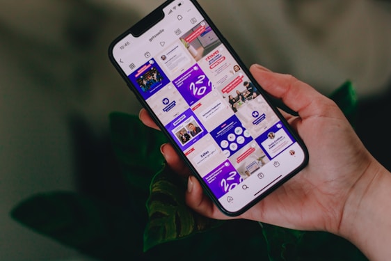 A hand holding a smartphone displaying an Instagram profile grid. The screen features various posts with vibrant colors and text, focusing on motivational content and social media strategies. The background includes leafy plants, adding a natural touch.