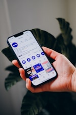 A hand holds a smartphone displaying a social media profile interface app, with text, icons, and images visible on the screen against a blurred plant background.