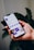 A hand holds a smartphone displaying a social media profile interface app, with text, icons, and images visible on the screen against a blurred plant background.
