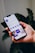 A hand holds a smartphone displaying a social media profile interface app, with text, icons, and images visible on the screen against a blurred plant background.