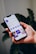 A hand holds a smartphone displaying a social media profile interface app, with text, icons, and images visible on the screen against a blurred plant background.