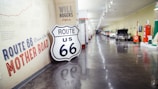 A hallway in a museum featuring a display dedicated to Route 66. The walls are adorned with vintage signage and historical information about the iconic highway. A prominent Route 66 shield sign is leaned against the wall. The corridor is lined with classic cars and exhibits related to the history of Route 66.