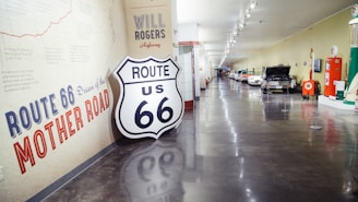 A hallway in a museum featuring a display dedicated to Route 66. The walls are adorned with vintage signage and historical information about the iconic highway. A prominent Route 66 shield sign is leaned against the wall. The corridor is lined with classic cars and exhibits related to the history of Route 66.