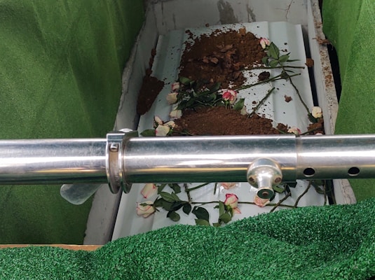 The image depicts a partially covered casket in a burial setting, surrounded by artificial green turf. There is a steel railing in the foreground. Some soil and a few pink and white roses are scattered over the casket.