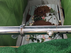 The image depicts a partially covered casket in a burial setting, surrounded by artificial green turf. There is a steel railing in the foreground. Some soil and a few pink and white roses are scattered over the casket.