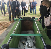 A group of people, dressed in formal clothing, stand around an open grave. The grave is surrounded by green fabric, and within it lies a coffin with roses placed on top. A woman in the foreground holds a single rose over the grave, while a man with a shovel stands nearby. The mood is somber, suggesting a funeral or memorial service.