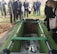 A group of people, dressed in formal clothing, stand around an open grave. The grave is surrounded by green fabric, and within it lies a coffin with roses placed on top. A woman in the foreground holds a single rose over the grave, while a man with a shovel stands nearby. The mood is somber, suggesting a funeral or memorial service.