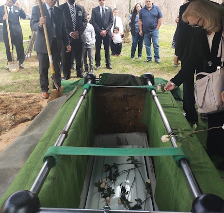 A group of people, dressed in formal clothing, stand around an open grave. The grave is surrounded by green fabric, and within it lies a coffin with roses placed on top. A woman in the foreground holds a single rose over the grave, while a man with a shovel stands nearby. The mood is somber, suggesting a funeral or memorial service.