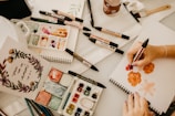 An artist is working on a painting using a variety of markers and watercolors. The workspace contains sketchpads, color palettes, brushes, and art markers spread across a table. A hand is holding a red ProMarker, creating floral designs on a blank page. A finished inspirational art piece with floral decorations is displayed on the left.