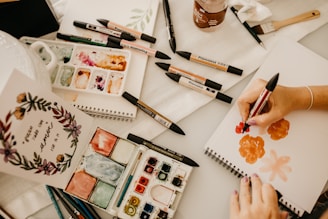 An artist is working on a painting using a variety of markers and watercolors. The workspace contains sketchpads, color palettes, brushes, and art markers spread across a table. A hand is holding a red ProMarker, creating floral designs on a blank page. A finished inspirational art piece with floral decorations is displayed on the left.