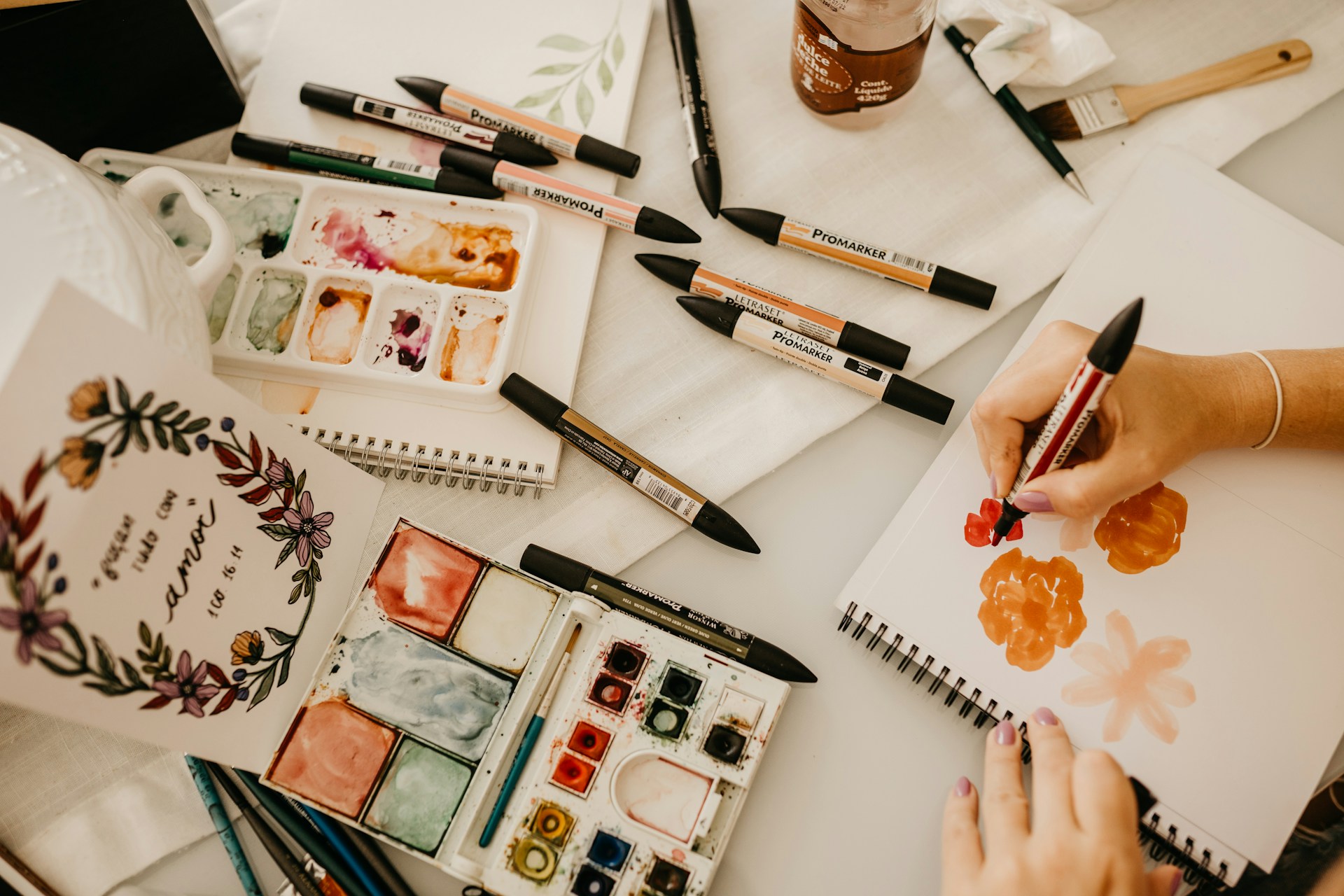 An artist is working on a painting using a variety of markers and watercolors. The workspace contains sketchpads, color palettes, brushes, and art markers spread across a table. A hand is holding a red ProMarker, creating floral designs on a blank page. A finished inspirational art piece with floral decorations is displayed on the left.