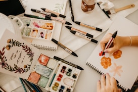 An artist is working on a painting using a variety of markers and watercolors. The workspace contains sketchpads, color palettes, brushes, and art markers spread across a table. A hand is holding a red ProMarker, creating floral designs on a blank page. A finished inspirational art piece with floral decorations is displayed on the left.