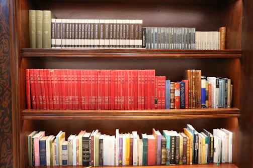 A wooden bookshelf filled with a variety of books arranged across three shelves. The top shelf contains a series of reference books with uniform covers in neutral tones. The middle shelf is dominated by a set of red encyclopedias, alongside a few smaller volumes in different colors. The bottom shelf holds a diverse collection of books, with varying colors and sizes.