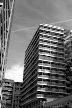 A black and white photograph of a modern high-rise residential building with numerous balconies, situated between other buildings. The perspective is from below, emphasizing the vertical lines and geometric structure. The sky is clear with some clouds.