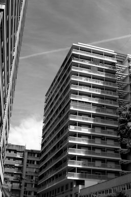 A black and white photograph of a modern high-rise residential building with numerous balconies, situated between other buildings. The perspective is from below, emphasizing the vertical lines and geometric structure. The sky is clear with some clouds.