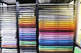 A neatly organized display of colored paper sheets stacked in metal shelves. The papers are arranged by color, ranging from whites and creams on the left to vibrant shades of red, orange, yellow, green, blue, and purple on the right. The shelves create a visually appealing gradient effect.