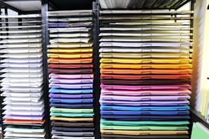 A neatly organized display of colored paper sheets stacked in metal shelves. The papers are arranged by color, ranging from whites and creams on the left to vibrant shades of red, orange, yellow, green, blue, and purple on the right. The shelves create a visually appealing gradient effect.