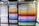 A neatly organized display of colored paper sheets stacked in metal shelves. The papers are arranged by color, ranging from whites and creams on the left to vibrant shades of red, orange, yellow, green, blue, and purple on the right. The shelves create a visually appealing gradient effect.