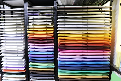 A neatly organized display of colored paper sheets stacked in metal shelves. The papers are arranged by color, ranging from whites and creams on the left to vibrant shades of red, orange, yellow, green, blue, and purple on the right. The shelves create a visually appealing gradient effect.