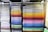 A neatly organized display of colored paper sheets stacked in metal shelves. The papers are arranged by color, ranging from whites and creams on the left to vibrant shades of red, orange, yellow, green, blue, and purple on the right. The shelves create a visually appealing gradient effect.