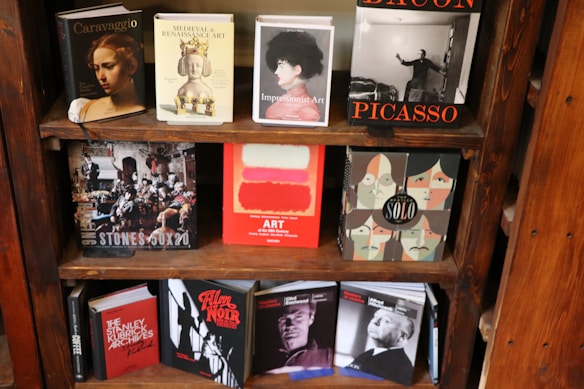A bookshelf displays a variety of art and film-related books. The top shelf includes titles on Caravaggio, Medieval and Renaissance Art, Impressionist Art, and a large book on Bacon and Picasso. The middle shelf has books on The Rolling Stones, contemporary art with abstract color blocks, and a graphic design with illustrated portraits. The bottom shelf features books on Stanley Kubrick, film noir, and Alfred Hitchcock.