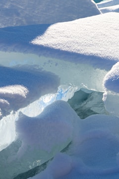 Chunks of ice and snow with a smooth, frosty texture are layered together. The ice appears translucent with a bluish hue, while the snow on top shows a bright white color. The layer of snow and ice captures sunlight, reflecting a soft glow.