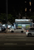 A nighttime urban scene with a newsstand labeled 'Banca Trianon Masp' in the center. Several cars are visible, driving past on a multi-lane road. Streetlights and trees are present in the background, creating a mix of natural and urban elements. The sky appears dark, emphasizing the artificial lighting.