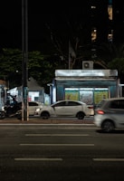 A nighttime urban scene with a newsstand labeled 'Banca Trianon Masp' in the center. Several cars are visible, driving past on a multi-lane road. Streetlights and trees are present in the background, creating a mix of natural and urban elements. The sky appears dark, emphasizing the artificial lighting.