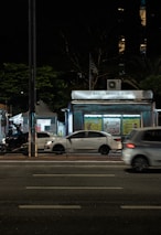 A nighttime urban scene with a newsstand labeled 'Banca Trianon Masp' in the center. Several cars are visible, driving past on a multi-lane road. Streetlights and trees are present in the background, creating a mix of natural and urban elements. The sky appears dark, emphasizing the artificial lighting.