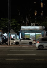 A nighttime urban scene with a newsstand labeled 'Banca Trianon Masp' in the center. Several cars are visible, driving past on a multi-lane road. Streetlights and trees are present in the background, creating a mix of natural and urban elements. The sky appears dark, emphasizing the artificial lighting.