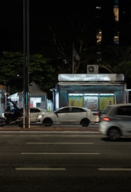 A nighttime urban scene with a newsstand labeled 'Banca Trianon Masp' in the center. Several cars are visible, driving past on a multi-lane road. Streetlights and trees are present in the background, creating a mix of natural and urban elements. The sky appears dark, emphasizing the artificial lighting.