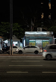 A nighttime urban scene with a newsstand labeled 'Banca Trianon Masp' in the center. Several cars are visible, driving past on a multi-lane road. Streetlights and trees are present in the background, creating a mix of natural and urban elements. The sky appears dark, emphasizing the artificial lighting.