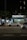 A nighttime urban scene with a newsstand labeled 'Banca Trianon Masp' in the center. Several cars are visible, driving past on a multi-lane road. Streetlights and trees are present in the background, creating a mix of natural and urban elements. The sky appears dark, emphasizing the artificial lighting.