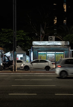 A nighttime urban scene with a newsstand labeled 'Banca Trianon Masp' in the center. Several cars are visible, driving past on a multi-lane road. Streetlights and trees are present in the background, creating a mix of natural and urban elements. The sky appears dark, emphasizing the artificial lighting.