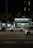 A nighttime urban scene with a newsstand labeled 'Banca Trianon Masp' in the center. Several cars are visible, driving past on a multi-lane road. Streetlights and trees are present in the background, creating a mix of natural and urban elements. The sky appears dark, emphasizing the artificial lighting.