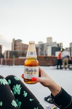 A close-up of a person's hand with bright red nails holding a bottle of Modelo beer. The background features an urban setting with blurred buildings and a few people standing, likely on a rooftop. The person appears to be wearing black clothing with green floral patterns and has a small cross tattoo on their wrist.