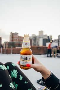A close-up of a person's hand with bright red nails holding a bottle of Modelo beer. The background features an urban setting with blurred buildings and a few people standing, likely on a rooftop. The person appears to be wearing black clothing with green floral patterns and has a small cross tattoo on their wrist.