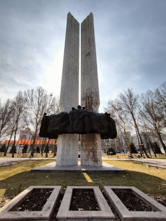 Tall concrete monument consisting of two vertical pillars with a dark sculpture of human figures draped over the structure. The monument is set in a park with barren trees and flags in the background, suggesting a public setting.