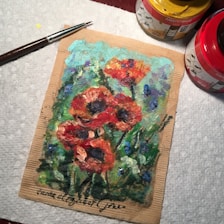 A vibrant painting of red poppies with dark centers, surrounded by green foliage and hints of blue flowers. The artwork is created on a piece of cardboard and is accompanied by a paintbrush and two jars of paint marked 'Amsterdam' in yellow and red respectively. The setting appears to be on a textured white surface, possibly a paper towel.