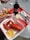 A culinary scene featuring a red tray with a container of freshly cooked lobster accompanied by a small cup of lemon butter and a slice of lemon. To the left, there is a basket of golden French fries in checkered paper. A bottle of Diet Coke, a small cup of coleslaw, and dining utensils including a fork, knife, and lobster crackers are also visible. The setting suggests a casual dining experience, possibly outdoors.