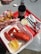 A culinary scene featuring a red tray with a container of freshly cooked lobster accompanied by a small cup of lemon butter and a slice of lemon. To the left, there is a basket of golden French fries in checkered paper. A bottle of Diet Coke, a small cup of coleslaw, and dining utensils including a fork, knife, and lobster crackers are also visible. The setting suggests a casual dining experience, possibly outdoors.